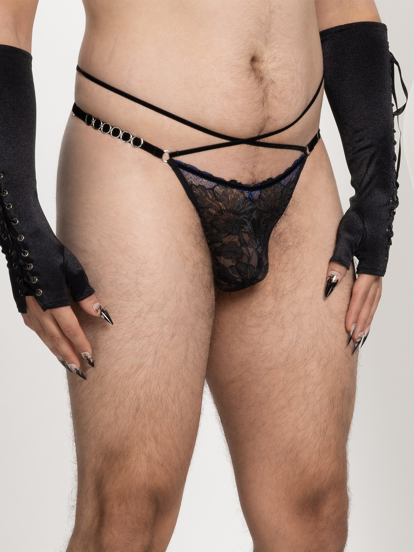 Model wearing a pouch fit thong made with floral black and blue lace, adjustable velvet straps and silver o-ring details on the hips, and 2 crossing straps on the waist.