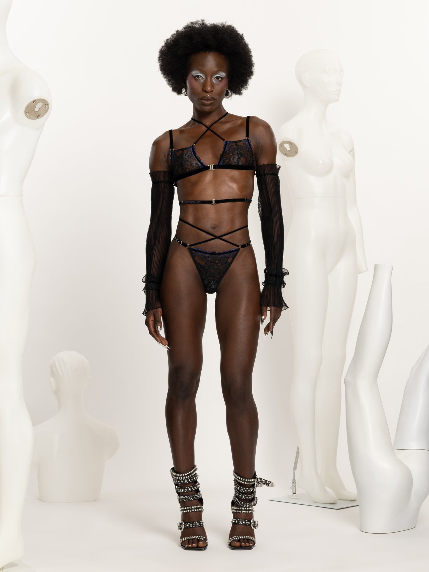 Full body view of a model wearing a black and blue lace lingerie set including bra and matching regular fit thong with black velvet straps across the chest waist and hips and silver o-rings details.