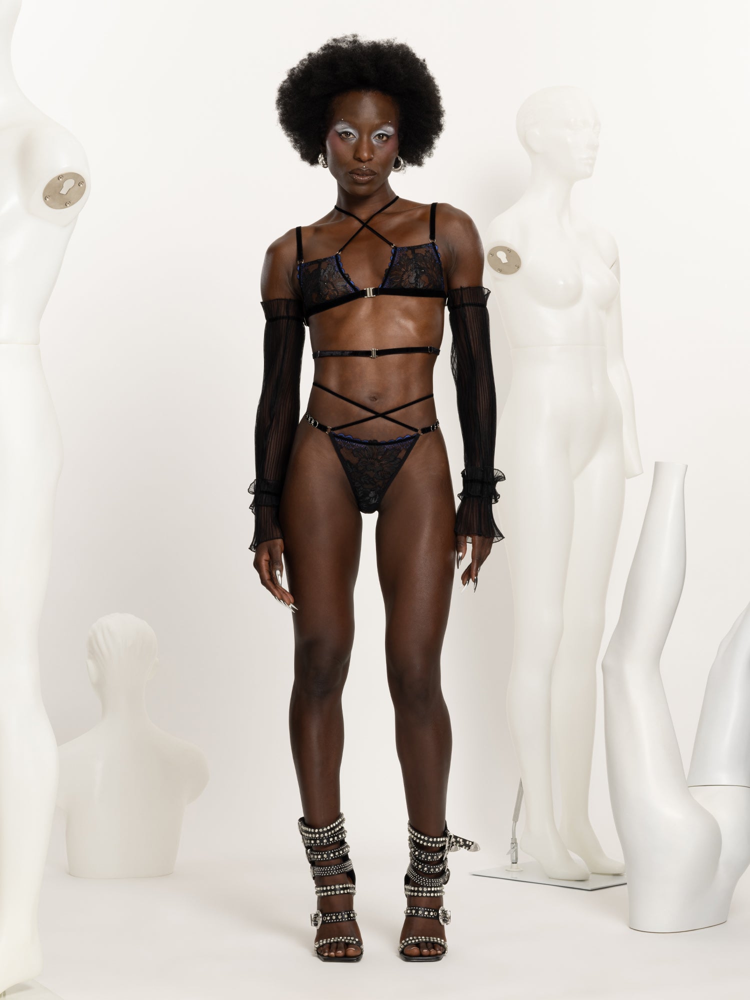 Full body view of a model wearing a black and blue lace lingerie set including bra and matching regular fit thong with black velvet straps across the chest waist and hips and silver o-rings details.