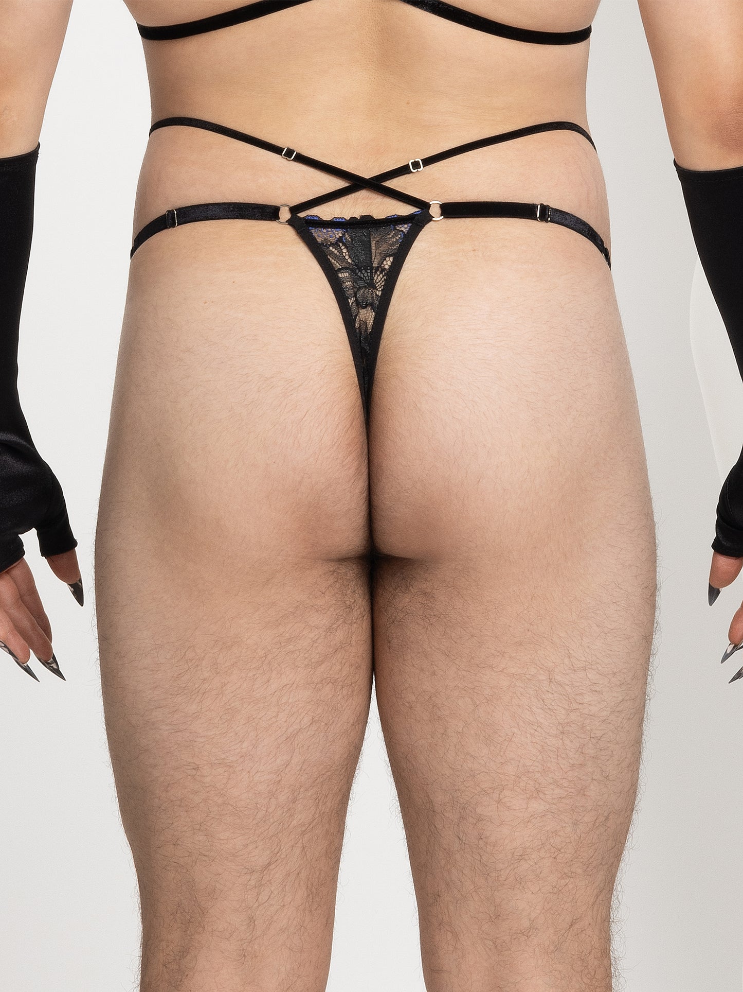 Back view of a model wearing pouch fit thong made with floral black and blue lace, adjustable velvet straps and silver o-ring details on the hips, and 2 crossing straps on the waist. 