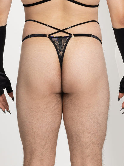 Back view of a model wearing pouch fit thong made with floral black and blue lace, adjustable velvet straps and silver o-ring details on the hips, and 2 crossing straps on the waist. 