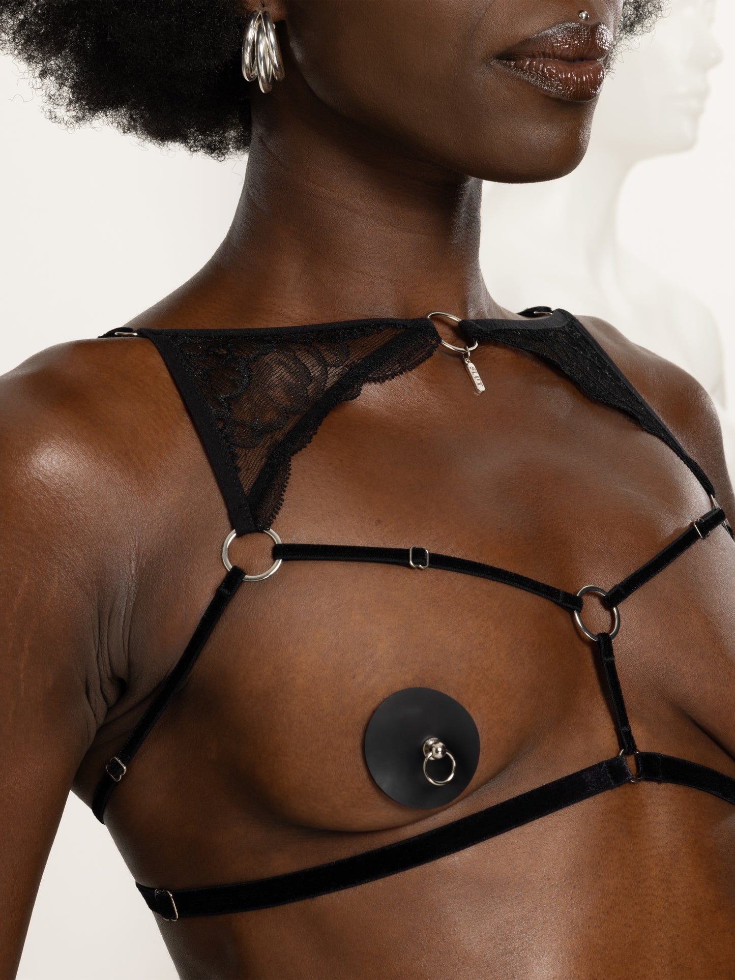 Close up view of an adjustable black velvet open cup harness with lace details above the chest, 'slut' engraved silver pendant and hardware.