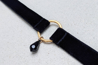 Close up view of an adjustable black velvet choker with a gold o-ring charm and a black crystal pendant.