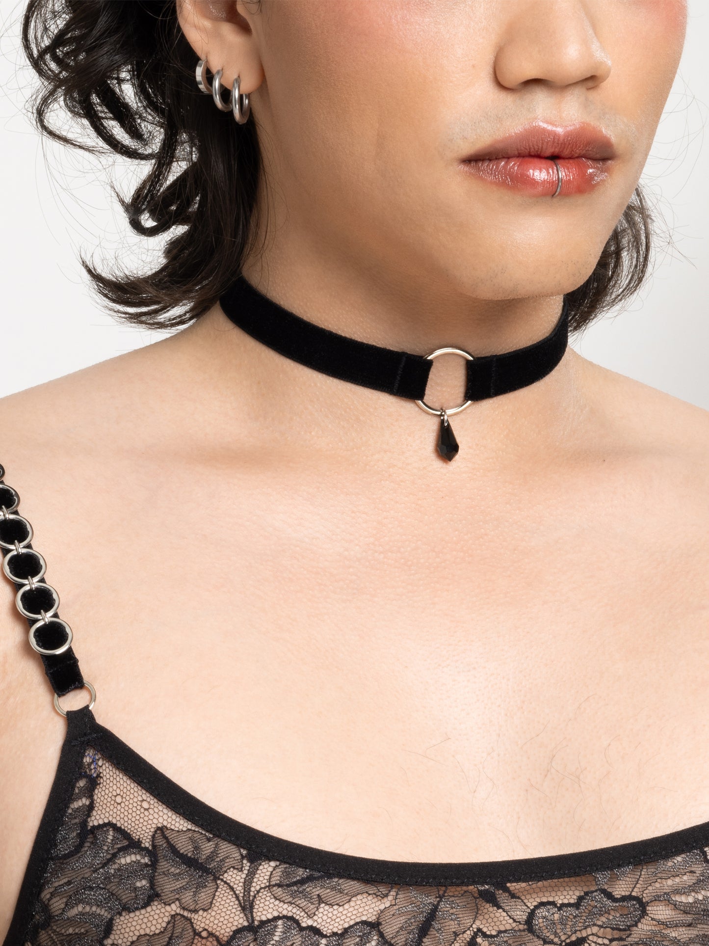 Close up view of a model wearing a black velvet choker made of a soft adjustable strap and a silver o-ring attached to a black crystal pendant. Also wearing a lace micro bralette.