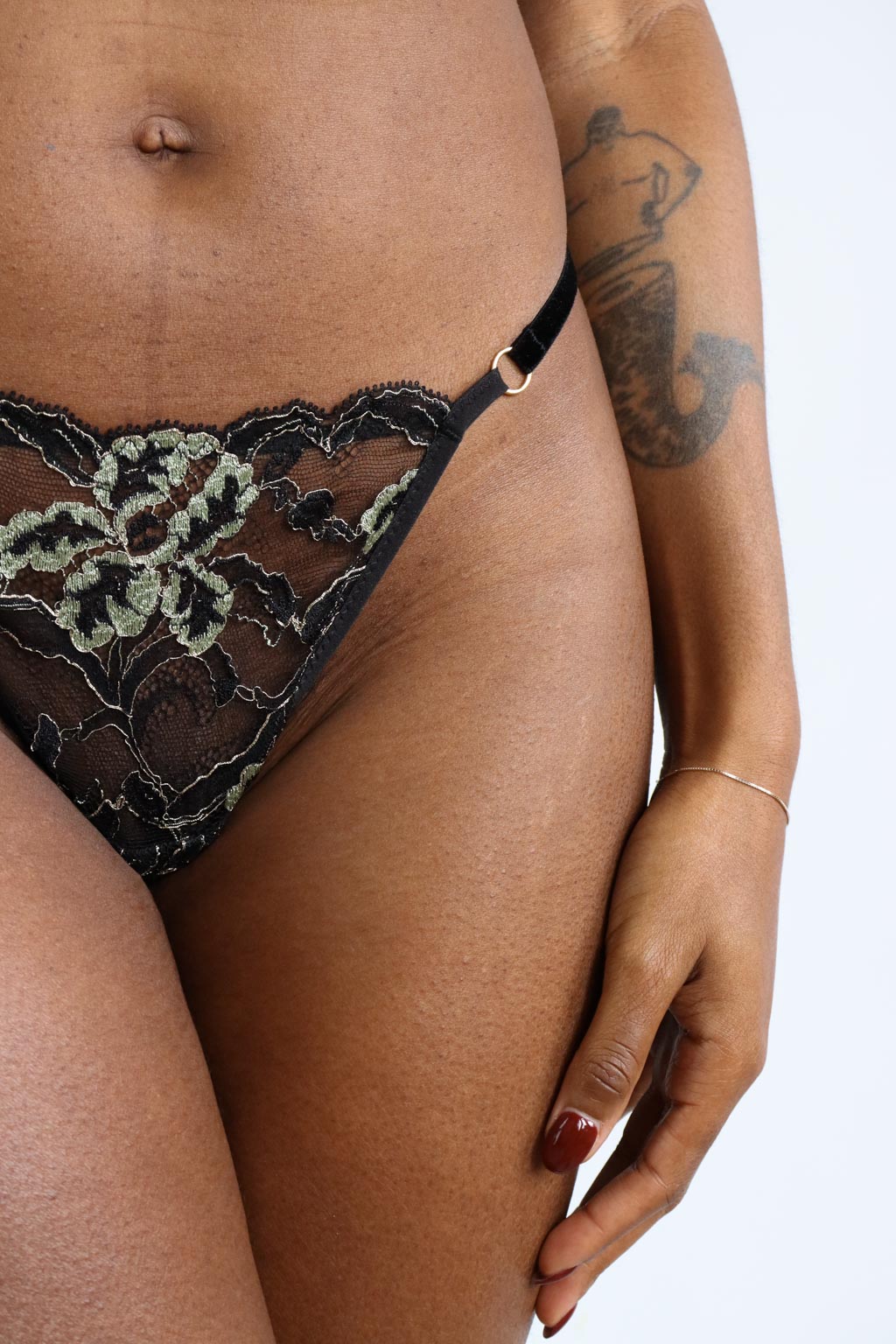 Close up view of a floral black and green lace thong with thin velvet straps and gold o-ring hardware.