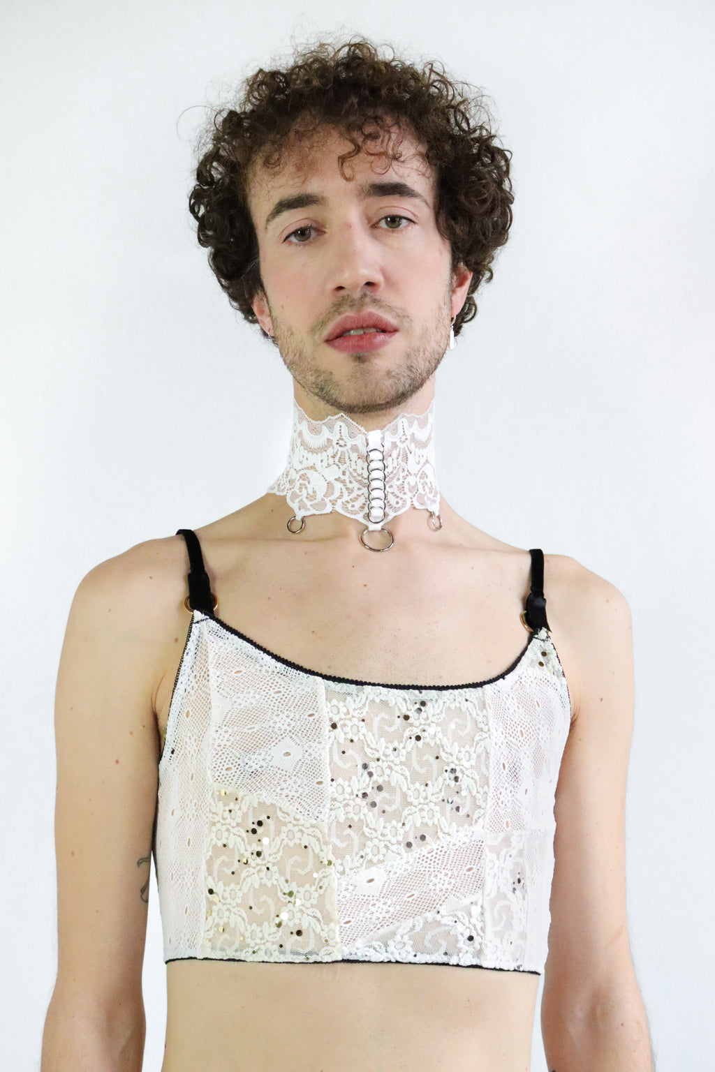 Bust view of a man wearing a white lace collar with silver o-ring pendants and a white adjustable lace crop top with velvet straps.