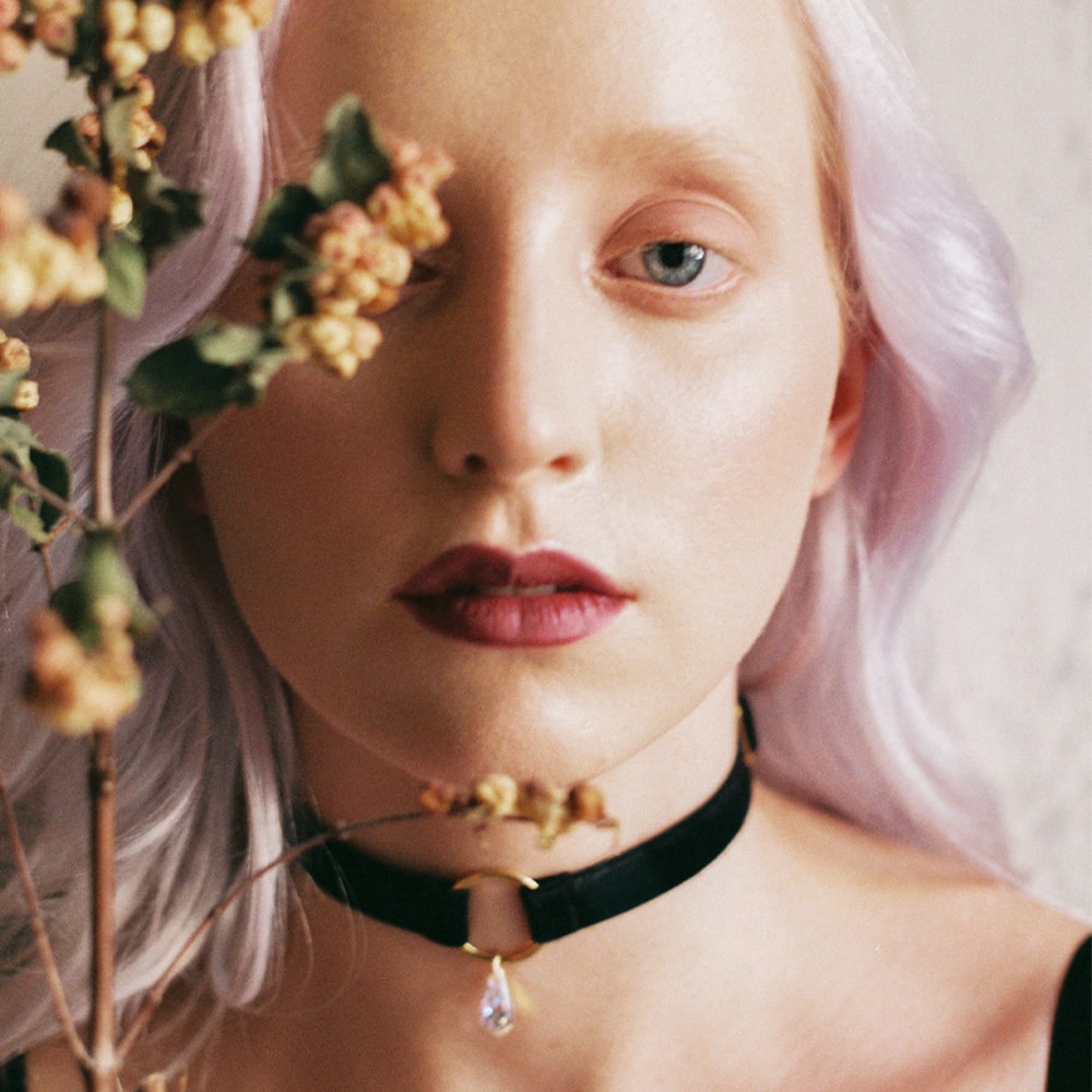Portait of a woman wearing a gold O-ring choker with black velvet straps and Swarovski crystal pendant. She is holding a dried flower in her hand, hiding partially her face.
