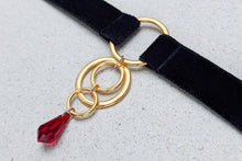 Load image into Gallery viewer, Close-up of an O-ring choker with black velvet straps, gold hardware and a red crystal pendant.
